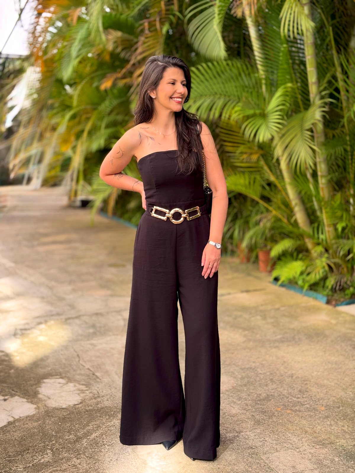 Dra. Jessica Motta, enfermeira esteta, em pé num caminho de jardim com vegetação tropical, de macacão preto e cinto dourado