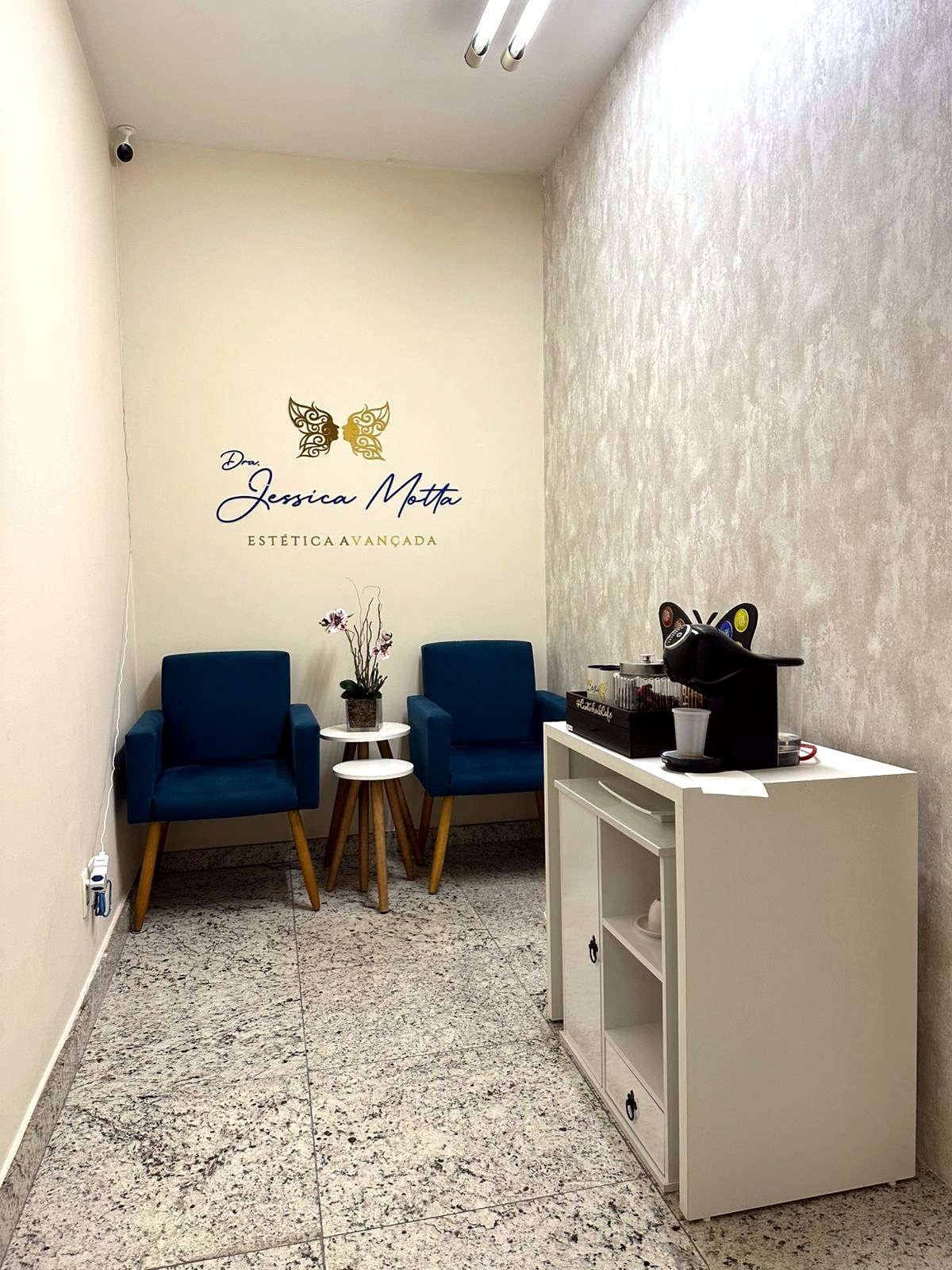 Sala do consultório da Dra. Jessica Motta, em Lourdes: duas poltronas de veludo azul, mesa lateral com flores secas e, na parede, a assinatura Estética Avançada com uma borboleta desenhada